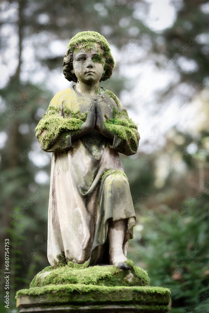 Small moss-covered cherub kneeling in prayer on the base of a gravestone