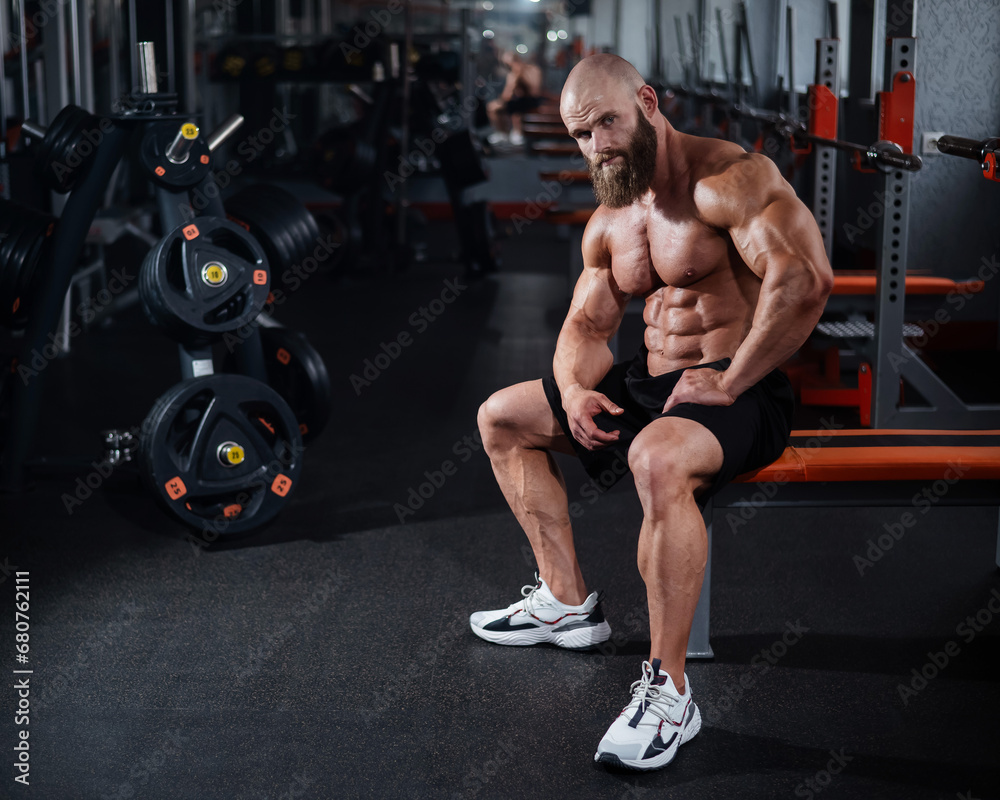 A muscular bald man in shorts is resting on a bench after a workout. Bodybuilder showing off his shape in the gym. A muscular bald man in shorts is resting on a bench after a workout. Bodybuilder showing off his shape in the gym.