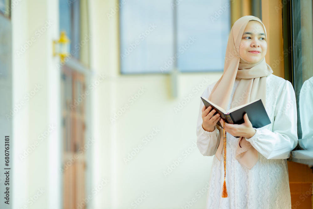 The image of an Asian Muslim woman in the Islamic religion in hijab in cream color. reading the Quran and having a happy Staying in a beautiful mosque, Arabic word translation: The Holy Al Quran.