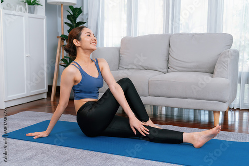 Asian woman in sportswear doing yoga exercise on fitness mat as her home workout training routine. Healthy body care and calm meditation in yoga lifestyle with comfortable and relaxation. Vigorous
