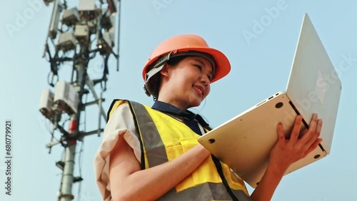 Asian female engineer wearing helmet performs field work telecommunications tower controlling mobile power installation using laptop maintain 5G network installed high rise building.