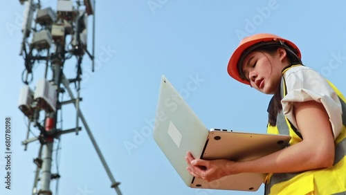 Asian female engineer wearing helmet performs field work telecommunications tower controlling mobile power installation using laptop maintain 5G network installed high rise building.