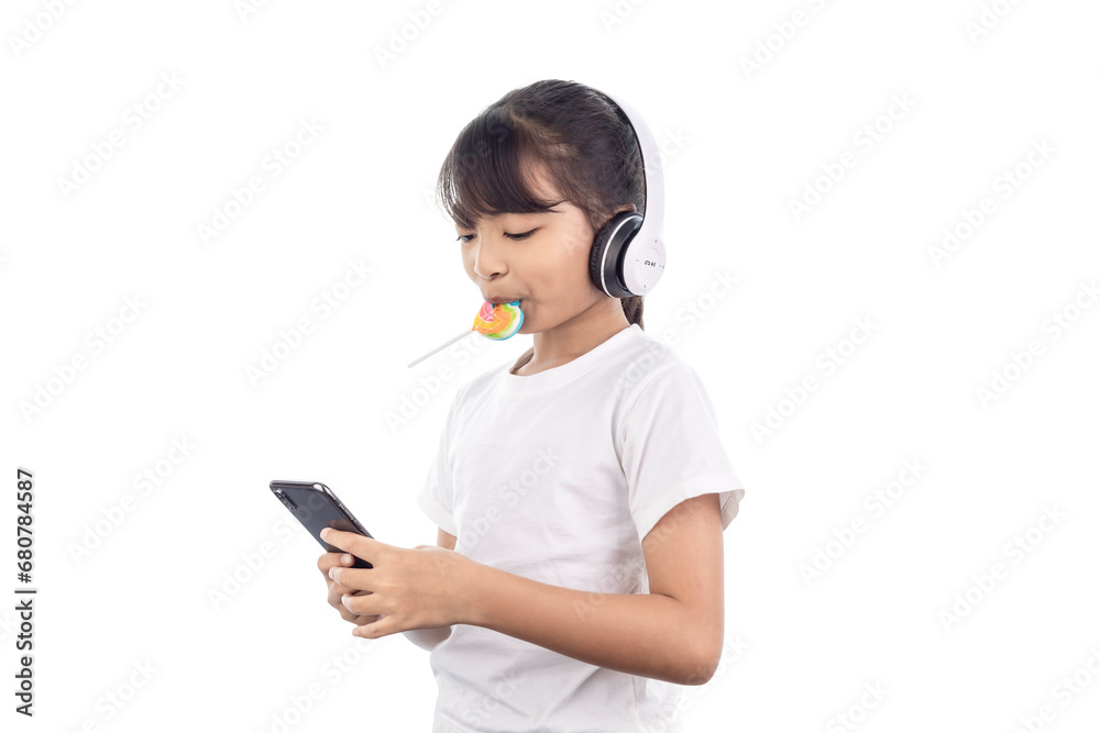 A happy Asian young girl playing music with her smartphone and wearing headphones and eating a lollipop isolated on white background with clipping path. A happy Asian young girl playing music with her smartphone and wearing headphones and eating a lollipop isolated on white background with clipping path.