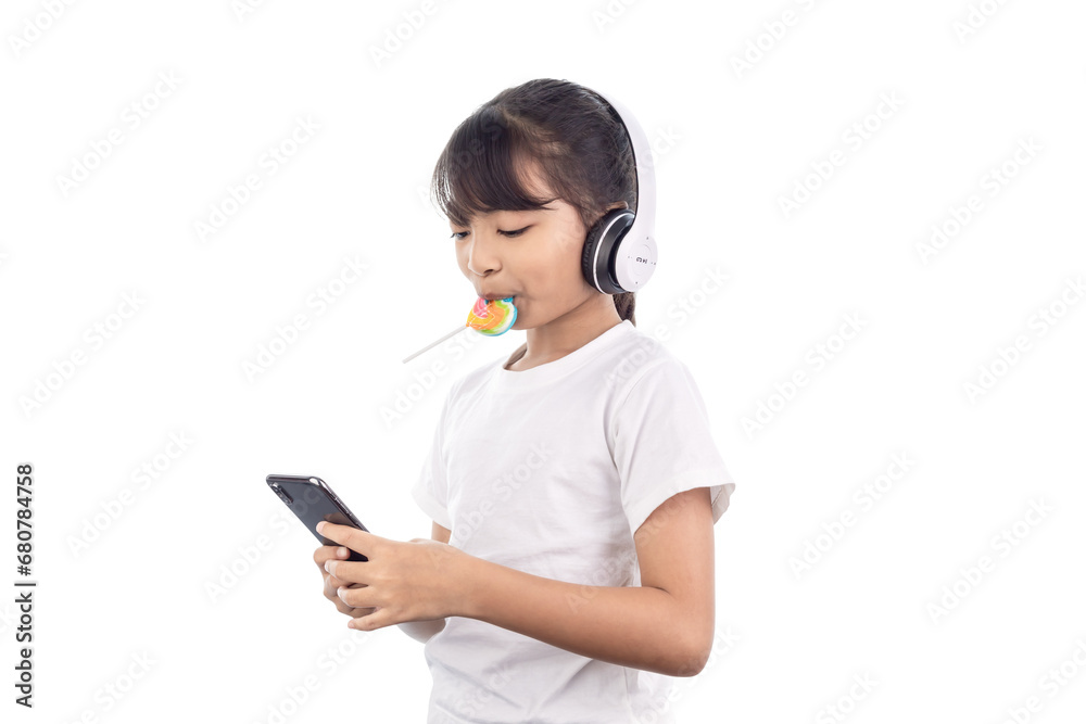 A happy Asian young girl playing music with her smartphone and wearing headphones and eating a lollipop isolated on white background with clipping path. A happy Asian young girl playing music with her smartphone and wearing headphones and eating a lollipop isolated on white background with clipping path.