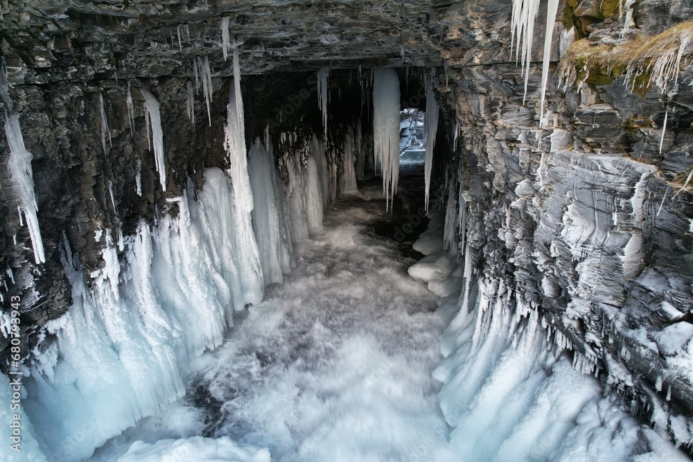 The drone hovers over the entrance of an ice cave in Björkliden, where water rushes beneath a stunning array of hanging icicles.