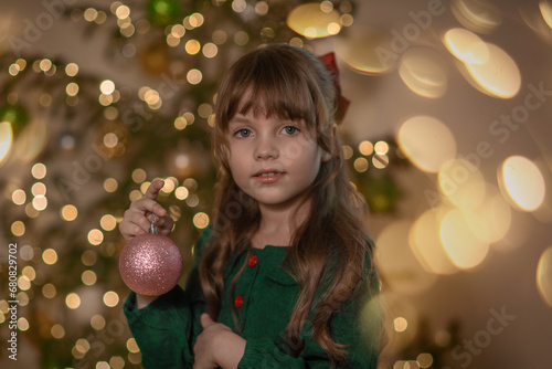 A portrait of a sweet beautiful smiling little 7-old girl wearing green dress, with red bow on the her brown long hair, decorate a Christmas tree with a pink shine glass ball at home at Christmas eve