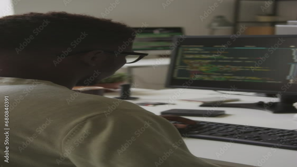 Vertical rear view waist up shot of African American guy in t-shirt and eyeglasses developing new software on desktop computer in office, writing programming codes via keyboard Vertical rear view waist up shot of African American guy in t-shirt and eyeglasses developing new software on desktop computer in office, writing programming codes via keyboard