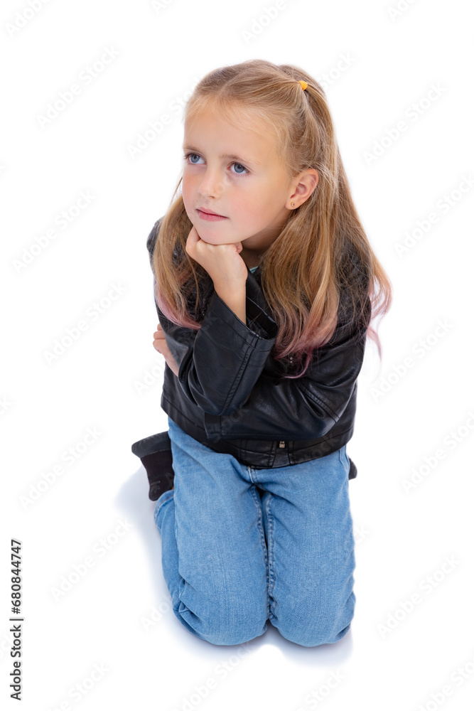 Serious seven years old girl sitting on the floor supported face with her fist, isolated on white background. Portrait of cute child in black leather jacket and blue jeans posing in studio. Serious seven years old girl sitting on the floor supported face with her fist, isolated on white background. Portrait of cute child in black leather jacket and blue jeans posing in studio.