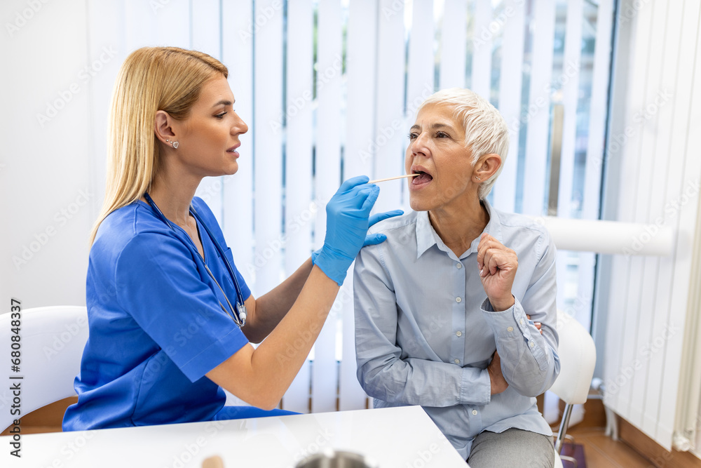 Obraz premium Senior patient opening her mouth for the doctor to look in her throat. Female doctor examining sore throat of patient in clinic. Otolaryngologist examines sore throat of patient.
