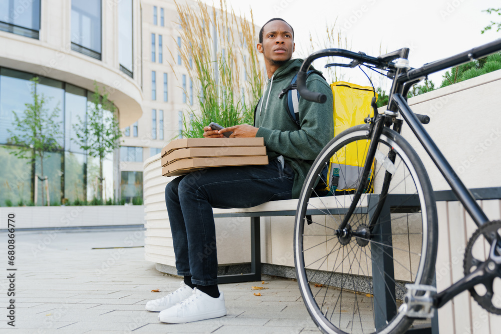 An African-American pizza deliveryman is sitting on a bench while waiting for customers. Timely delivery. An African-American pizza deliveryman is sitting on a bench while waiting for customers. Timely delivery.