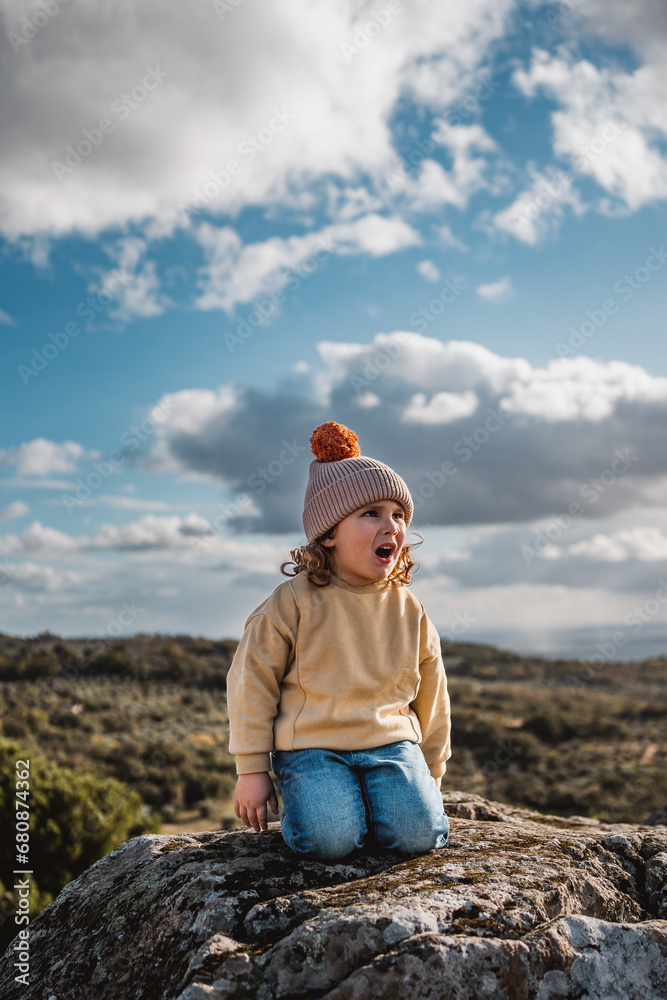 fashionable little boy with hat playing and happy, screaming excitedly, on top of a rock, in a stone castle with panoramic view of the mountains, on sunny day and with sky and forest background