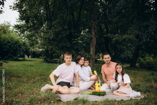 Fototapeta Naklejka Na Ścianę i Meble -  Happy big family with kids spending time together in field. Large company people eating on picnic in park. Mother, father, daughter, son sitting on blanket at sunset. Family holiday outdoors.