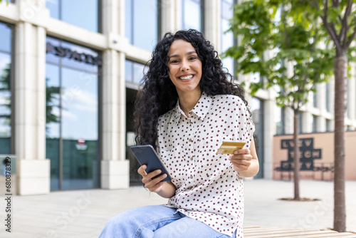 Portrait young beautiful woman with curly hair, sitting on a bench outside an office building, smiling and looking at camera, holding a phone, bank credit card, ordering online services, shopping.