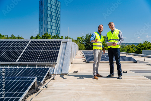 Two workers in safety vests discussing on the rooftop of a solar-powered company building while holding a tablet photo