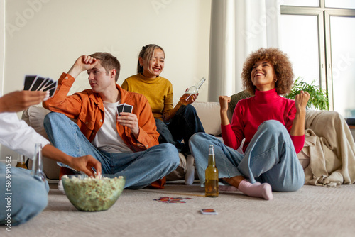 multiracial group of young friends playing cards drinking beer and having fun at home, students sitting on the floor