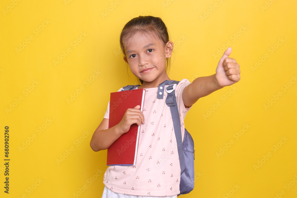 Adorable little girl holding book while carrying rucksack showing thumb up to camera with happy expression Adorable little girl holding book while carrying rucksack showing thumb up to camera with happy expression