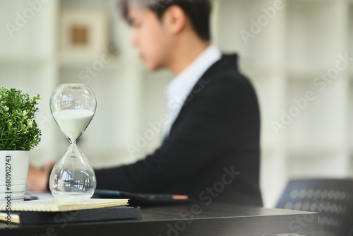 Hourglass on wooden office table with blurred businessman working on background. Time management concept photo