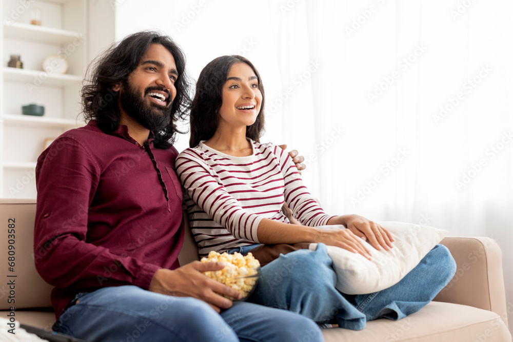 Positive young indian couple relaxing on sofa, watching TV,