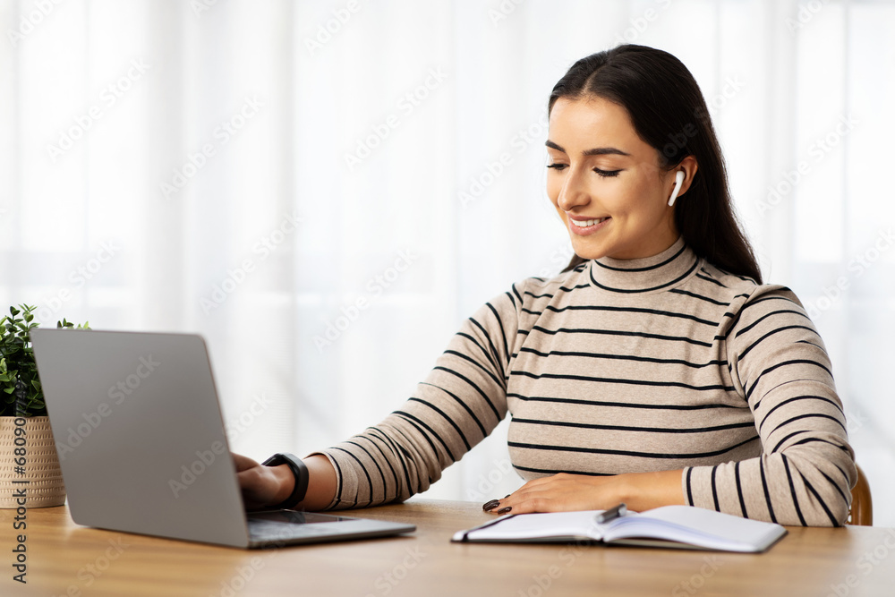 Positive young arab lady manager work at computer with wireless headphones, make notes Positive young arab lady manager work at computer with wireless headphones, make notes