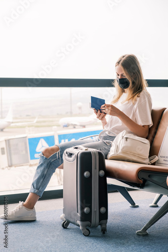 A young teenage girl is sitting at the airport waiting for her flight next to her suitcase. Girl holding a passport in her hands and wearing a mask