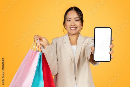 Happy millennial japanese woman with colorful bags, with purchases show phone with empty screen