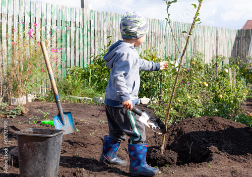 A little boy helps to dig a hole with his toy shovel to plant a young tree on a small rural plot on an autumn sunny day.