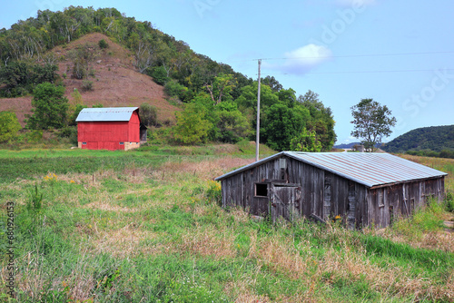 A ramshackle storage shed sits in a field near a bright red barn on a Midwestern farm. Behind the farm is a tree-covered hill. Green grass, late summer colors, brown dirt, blue partly-cloudy sky. photo