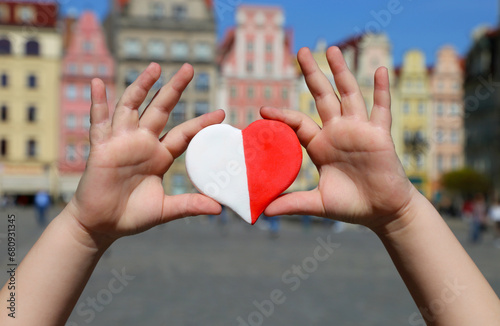 A red-white heart in the colors of the national flag of Poland in the hands of a child against the backdrop of the old town square. Independence Day of Poland. Freedom and Democracy