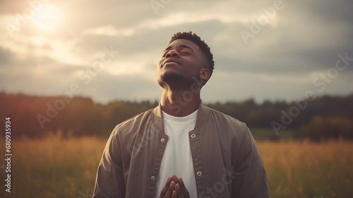 Stampa su tela A young black African man, spiritual and reflective, praying in a sunlit field, gazing upwards, captured with natural lighting and desaturated tones