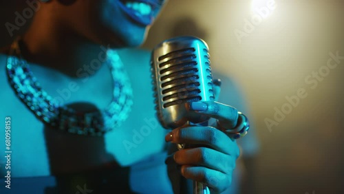 Medium closeup of attractive young Black woman singing blues in retro microphone while performing life on stage with blue light of projector on her face photo