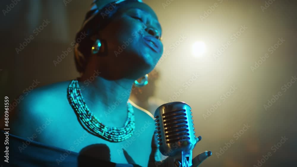 Chest up of artistic young Black female jazz vocalist wearing vintage beads necklace and patterned headscarf singing in retro microphone in blue light of projector