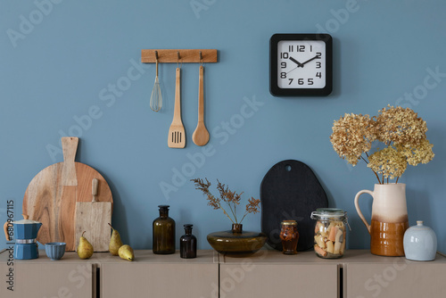 Interior design of kitchen interior with beige shelf, wooden cutboard, glass bottle, vase with flowers, vase with dried flowers, clock and personal accessories. Home decor. Template.