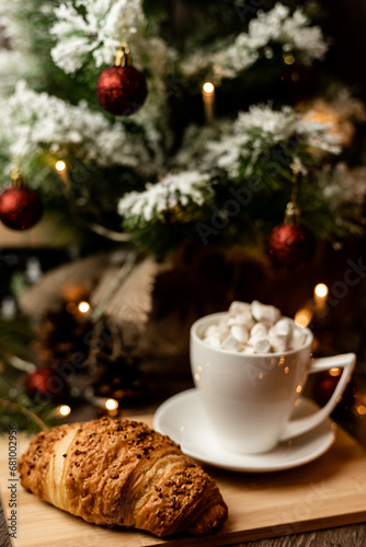 A marshmallow in a cup and croissant stand on the table near the Christmas tree