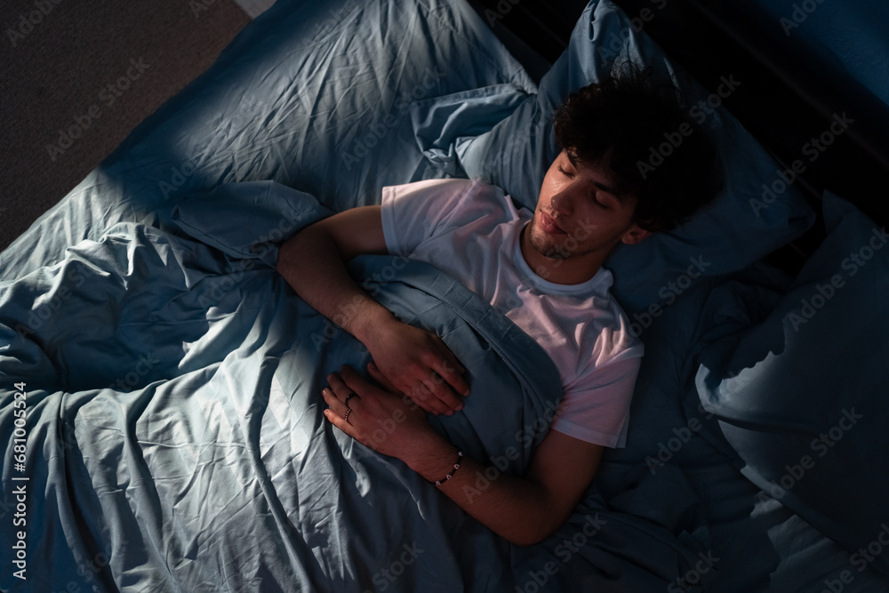 Top View of Arabic young man sleeping on a bed in his bedroom at night. Blue nightly light shining through the window. Top View of Arabic young man sleeping on a bed in his bedroom at night. Blue nightly light shining through the window.