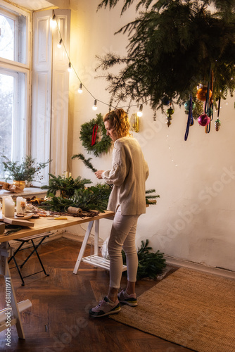 Vertical photo of young woman making handmade Christmas wreath at home from natural materials. DIY home decoration for New Year holidays. Blogger writes DIY instructions for social media. Scandinavian
