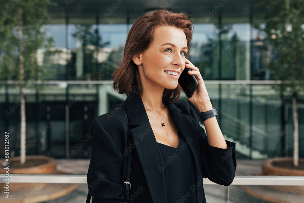 Smiling middle-aged woman talking on mobile phone at business building. Cheerful female with ginger hair in formal clothes standing outdoors and talking on her cell phone. Smiling middle-aged woman talking on mobile phone at business building. Cheerful female with ginger hair in formal clothes standing outdoors and talking on her cell phone.