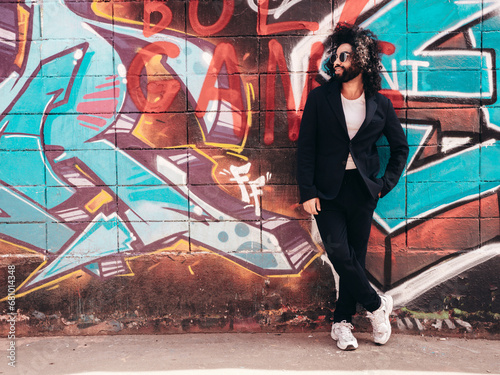 Handsome hipster model. Arabian man dressed in suit jacket clothes. Fashion male with long curly hairstyle posing in street near graffiti wall