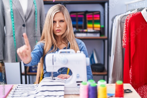 Blonde woman dressmaker designer using sew machine pointing with finger up and angry expression, showing no gesture