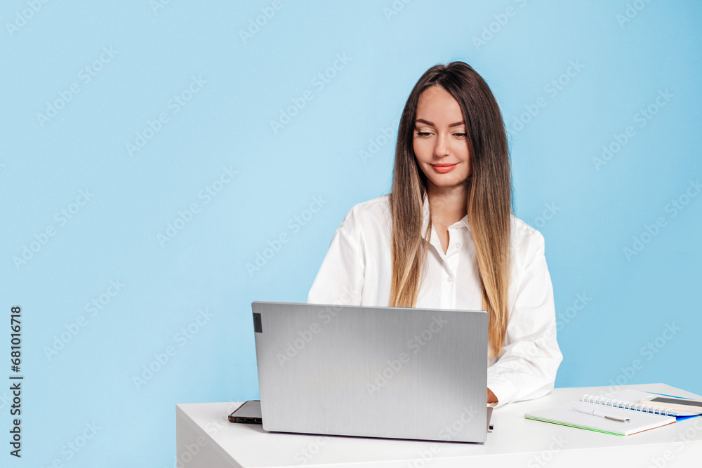 caucasian young business lady sitting at table with laptop and phone isolated over blue background in studio caucasian young business lady sitting at table with laptop and phone isolated over blue background in studio