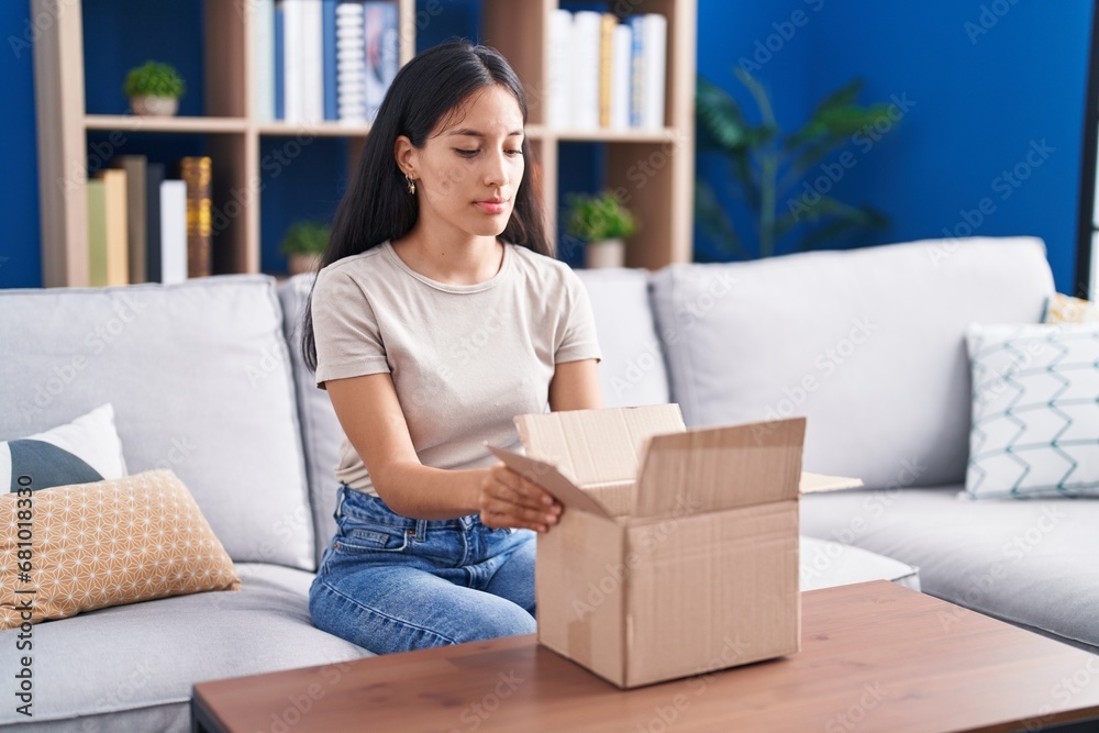 Young hispanic woman opening cardboard box relaxed with serious expression on face. simple and natural looking at the camera. Young hispanic woman opening cardboard box relaxed with serious expression on face. simple and natural looking at the camera.