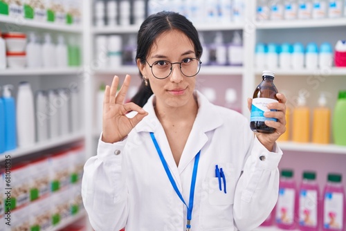 Young hispanic woman working at pharmacy drugstore holding syrup doing ok sign with fingers, smiling friendly gesturing excellent symbol