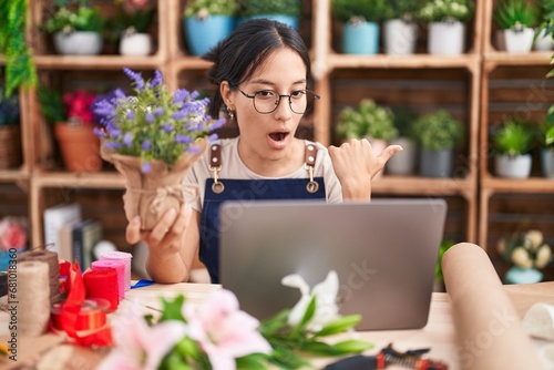Young hispanic woman working at florist shop doing video call surprised pointing with hand finger to the side, open mouth amazed expression. © Krakenimages.com