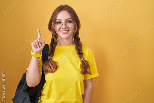 Young caucasian woman wearing student backpack over yellow background showing and pointing up with finger number one while smiling confident and happy.