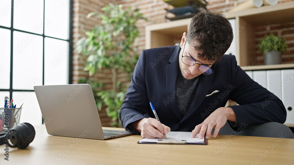Young hispanic man business worker using laptop writing on clipboard at office
