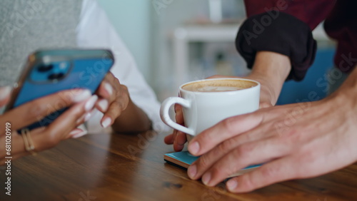 Barista putting cup coffee on table to woman sitting with smartphone closeup.