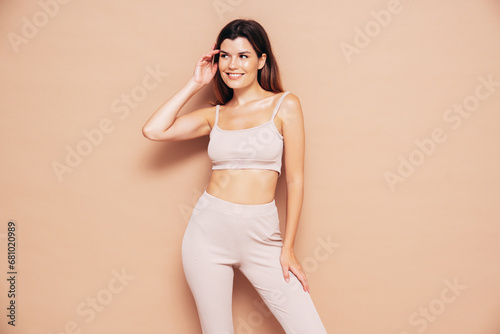 Young beautiful smiling female in trendy summer sport leggings and top clothes. Carefree woman posing near beige wall in studio. Positive model having fun indoors. Cheerful and happy © halayalex