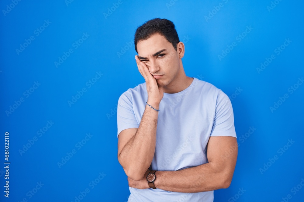 Young hispanic man standing over blue background thinking looking tired and bored with depression problems with crossed arms. Young hispanic man standing over blue background thinking looking tired and bored with depression problems with crossed arms.