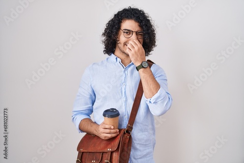 Hispanic man with curly hair drinking a cup of take away coffee smelling something stinky and disgusting, intolerable smell, holding breath with fingers on nose. bad smell