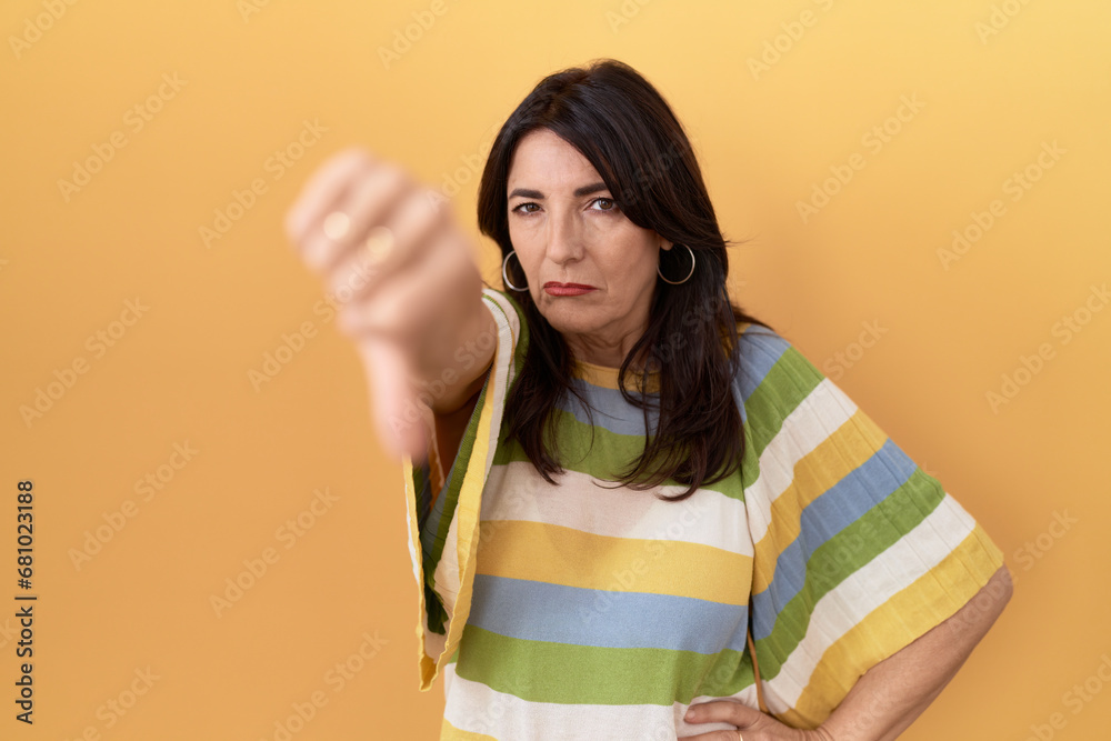Middle age hispanic woman standing over yellow background looking unhappy and angry showing rejection and negative with thumbs down gesture. bad expression. Middle age hispanic woman standing over yellow background looking unhappy and angry showing rejection and negative with thumbs down gesture. bad expression.