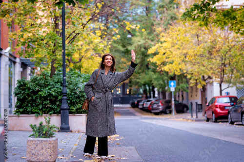 Happy mid aged woman with brunette hair walking on the city street and waving down a taxi with her hand © sepy
